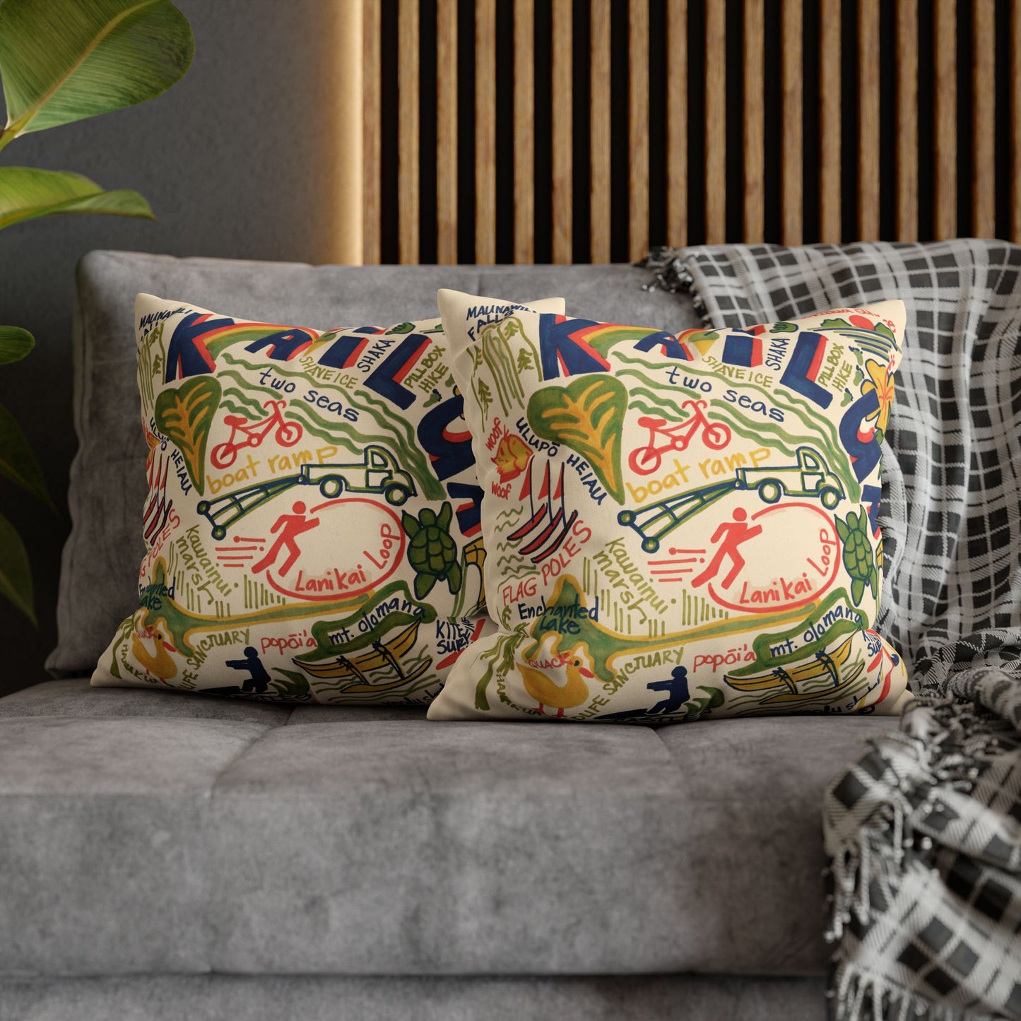 Two decorative throw pillows with a colorful vintage-style map print featuring bicycles, pedestrians, and text of Kailua, Oahu styled on a gray sofa with a wooden slat wall and a plaid blanket in the background.
