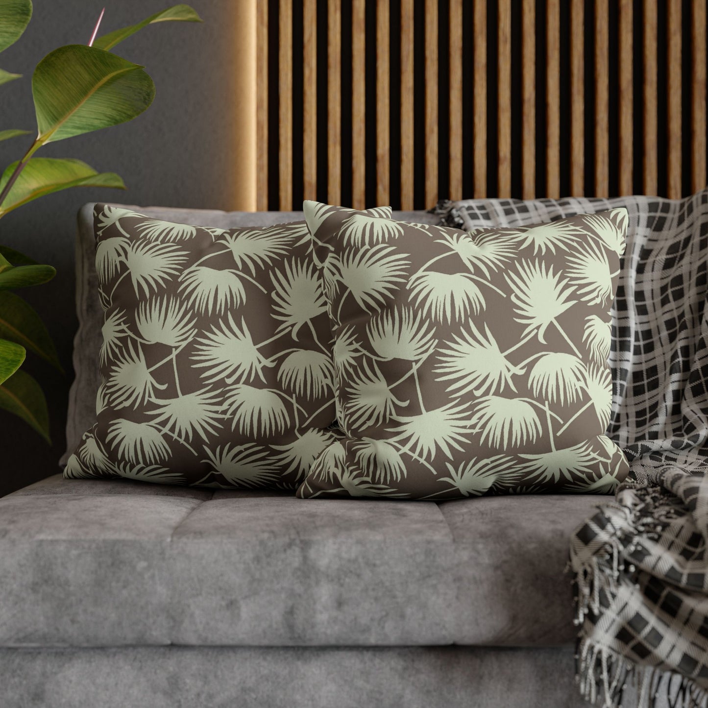 Two square throw pillows with a palm leaf pattern in olive green and taupe, styled on a gray sofa with a plaid throw blanket and a wooden slat wall background.