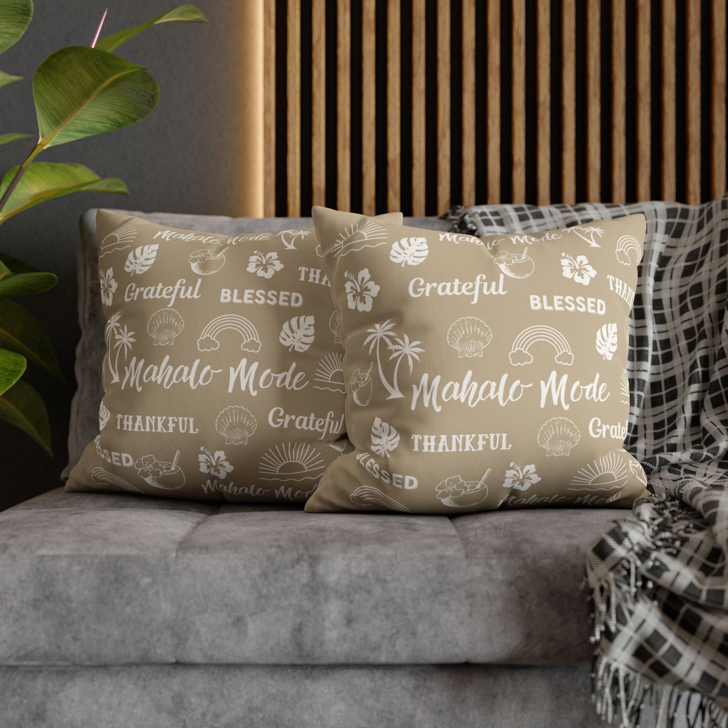 Two beige throw pillows with white tropical-themed printed text and icons, styled on a gray sofa with a plaid throw blanket and a wooden slat wall background.