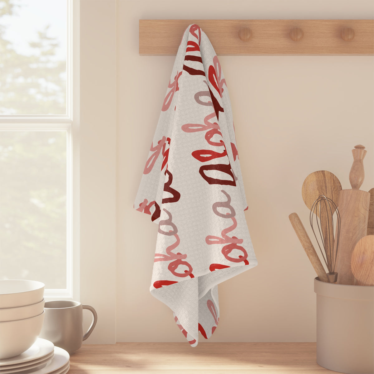Red Aloha Hawaiʻi kitchen towel with repeating script design in shades of red on white hanging off a wooden peg in bright kitchen