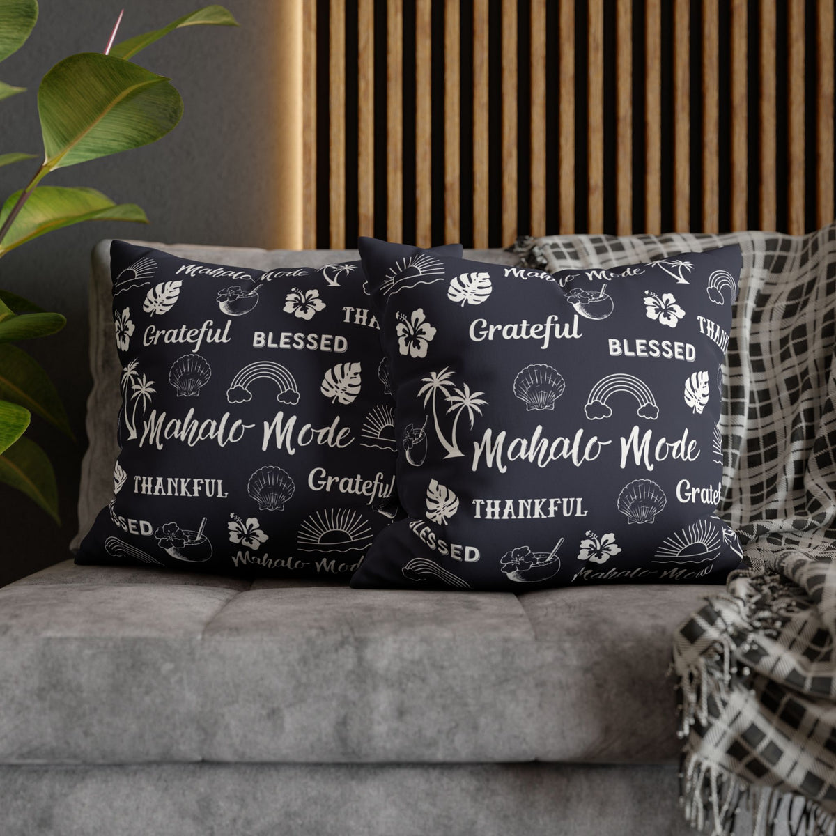 Two navy blue throw pillows with white tropical-themed text and illustrations, styled on a gray sofa with a plaid throw blanket and a wooden slat wall background.