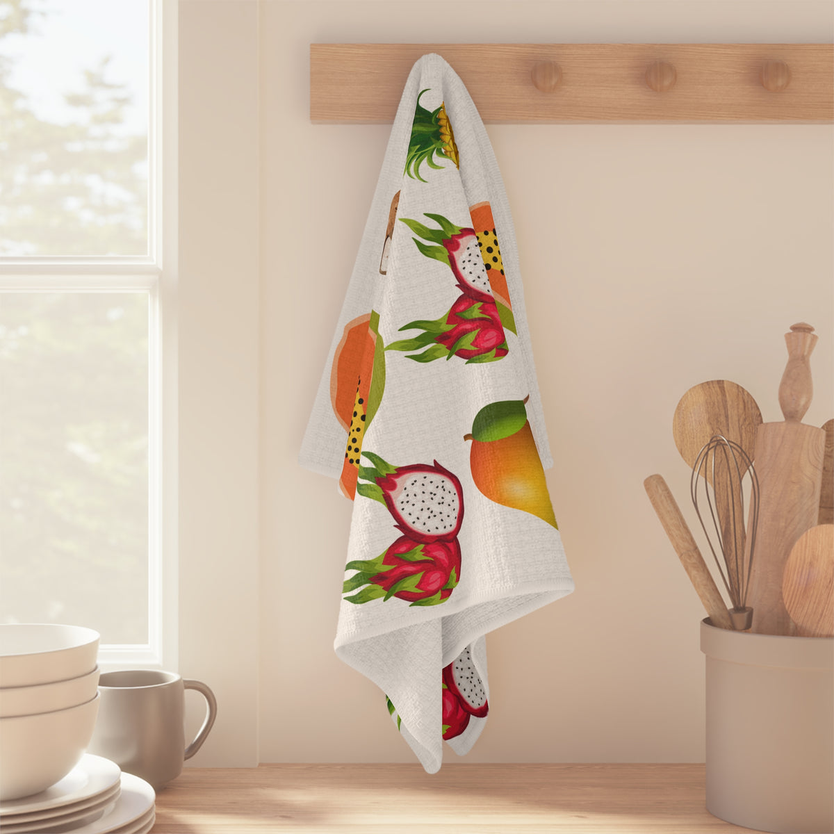 A white kitchen towel with a colorful fruit print hangs from a wooden peg on a wall in a bright kitchen setting.