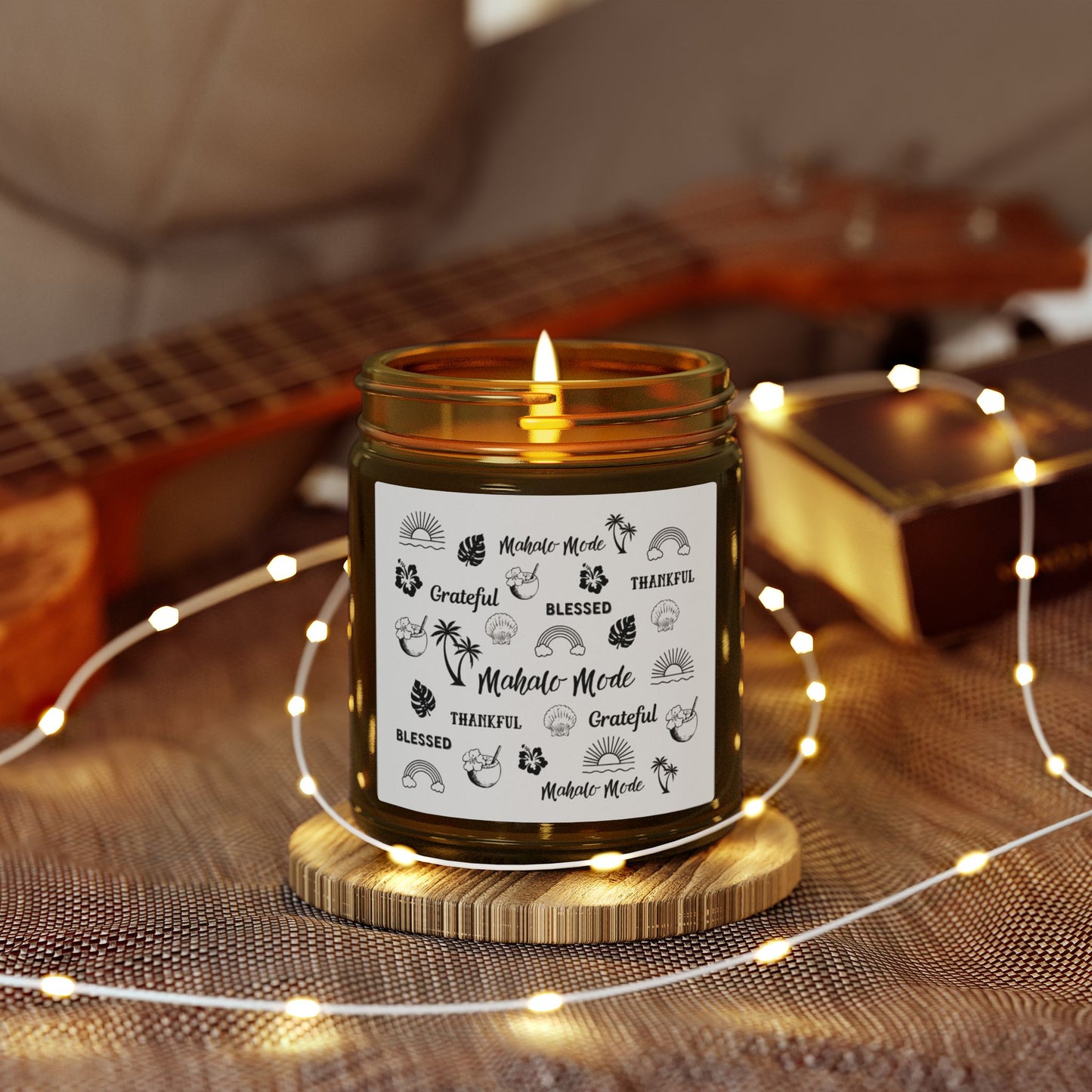 A dark amber glass jar candle with a gold metal screw-on lid and a white paper label featuring black hand-drawn style illustrations and the words 'Mahalo • Mode', 'Grateful', 'BLESSED', and 'THANKFUL'.on a wooden coaster, surrounded by string lights and a guitar in the background.