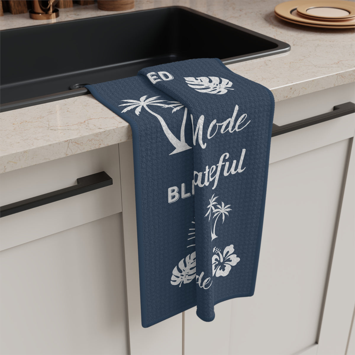 A navy blue kitchen towel with white tropical and text graphics hangs over the edge of a black sink in a modern kitchen setting.