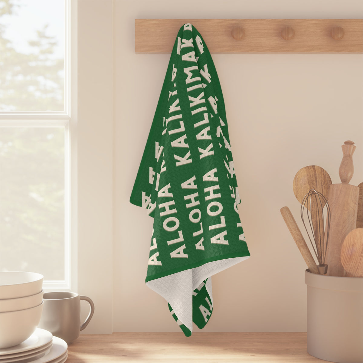 Green Aloha Kalikimaka holiday kitchen towel with repeating cream lettering hanging off a wood peg in kitchen