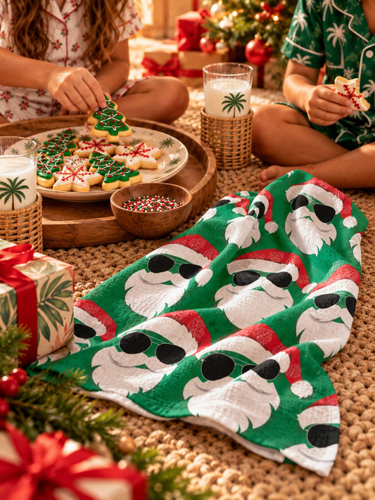 Green Santa kitchen towel with sunglasses draped on a kitchen counter beside Christmas cookies and a holiday mug.