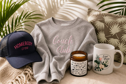 couch club sweatshirt, homebody crew trucker hat, mahalo mode candle and live pono tropical mug in a tropical living room