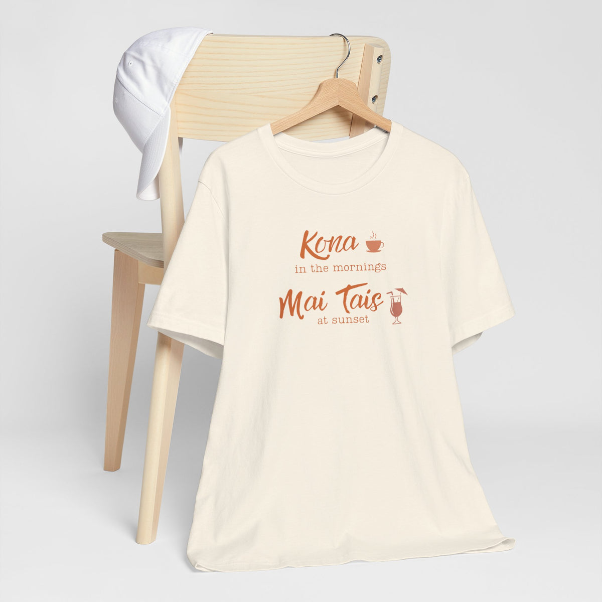 An ivory graphic t-shirt hanging on a wooden hanger, styled in front of a light wood chair with a white towel draped over the backrest. The shirt features a coral orange graphic that reads 'Kona in the mornings Mai Tais at sunset' with small coffee and cocktail illustrations.