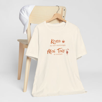 An ivory graphic t-shirt hanging on a wooden hanger, styled in front of a light wood chair with a white towel draped over the backrest. The shirt features a coral orange graphic that reads 'Kona in the mornings Mai Tais at sunset' with small coffee and cocktail illustrations.