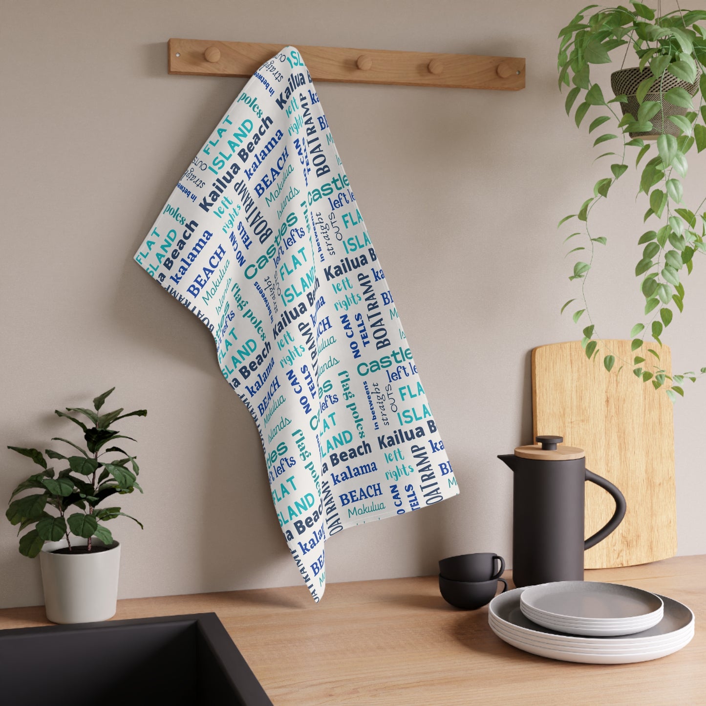 A white kitchen towel with a blue and aqua 'Kailua Surf Breaks' text pattern hangs from a wooden peg on a kitchen wall, styled with a black coffee pot, plates, and potted plants.