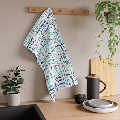 A white kitchen towel with a blue and aqua 'Kailua Surf Breaks' text pattern hangs from a wooden peg on a kitchen wall, styled with a black coffee pot, plates, and potted plants.