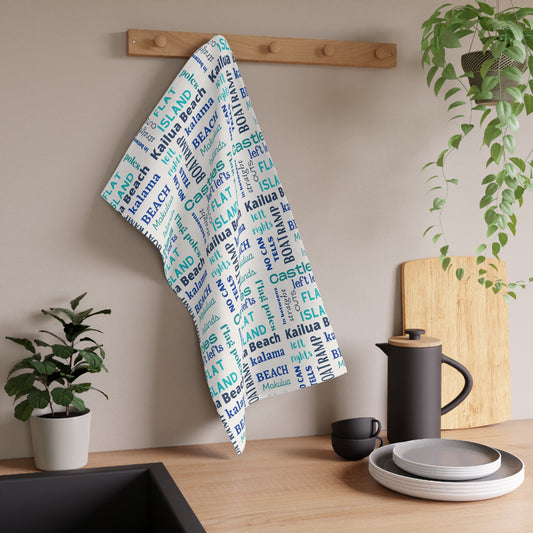 A white kitchen towel with a blue and aqua 'Kailua Surf Breaks' text pattern hangs from a wooden peg on a kitchen wall, styled with a black coffee pot, plates, and potted plants.