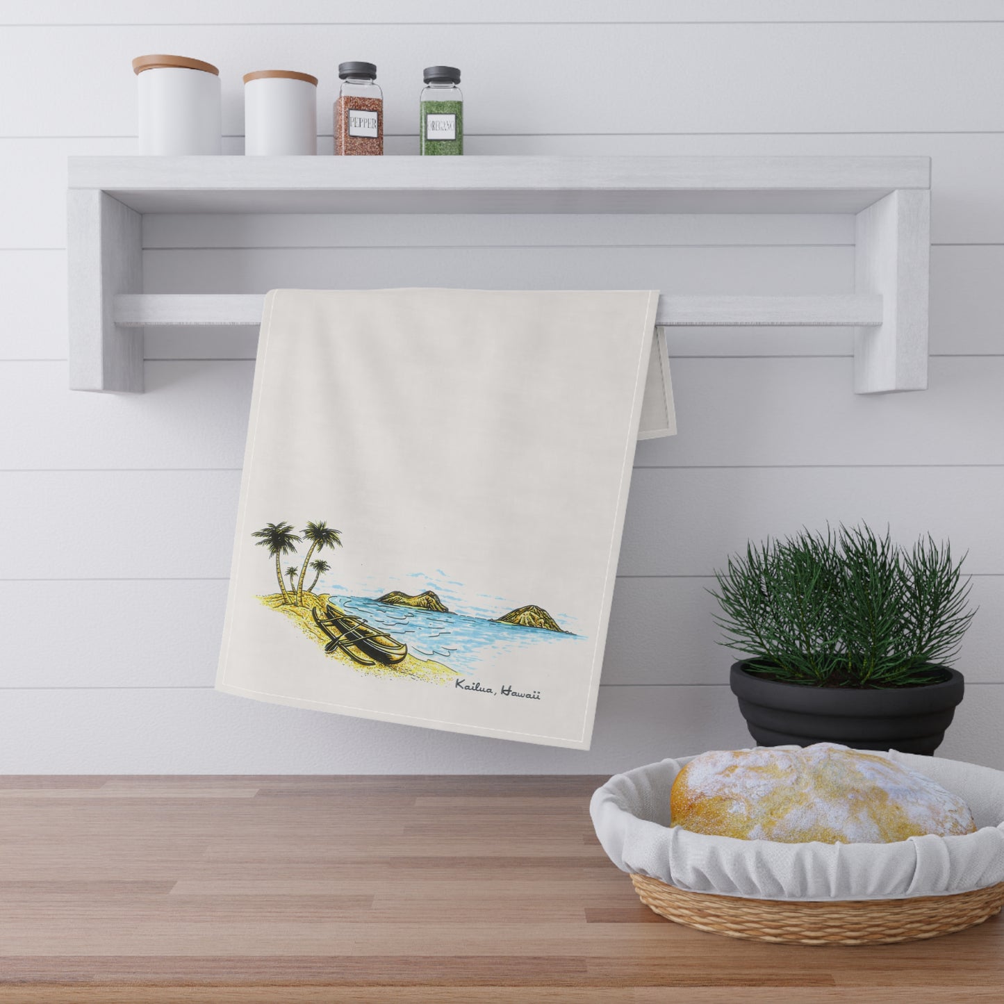 An off-white kitchen towel with a colorful beach illustration hanging from a white wall-mounted rack. The towel features a tropical scene with palm trees, a boat, and blue water, labeled 'Kailua, Hawaii'.