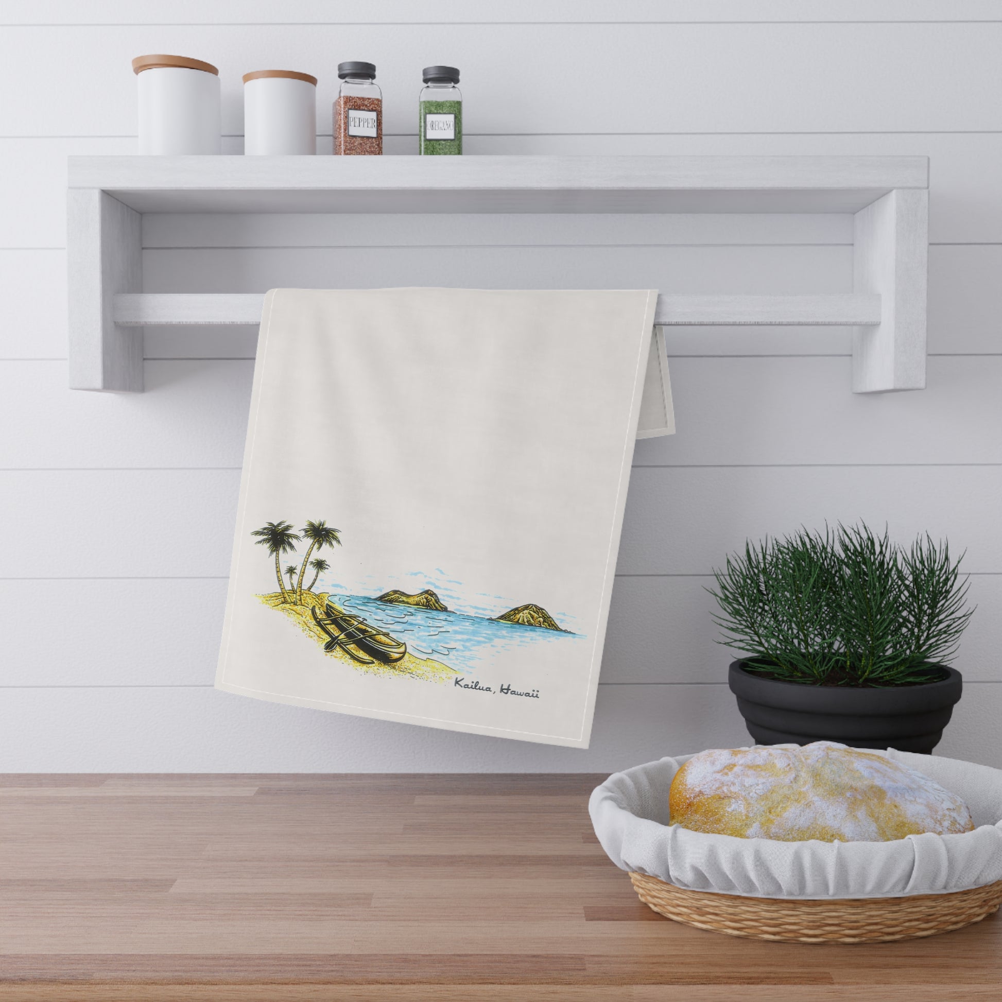 An off-white kitchen towel with a colorful beach illustration hanging from a white wall-mounted rack. The towel features a tropical scene with palm trees, a boat, and blue water, labeled 'Kailua, Hawaii'.