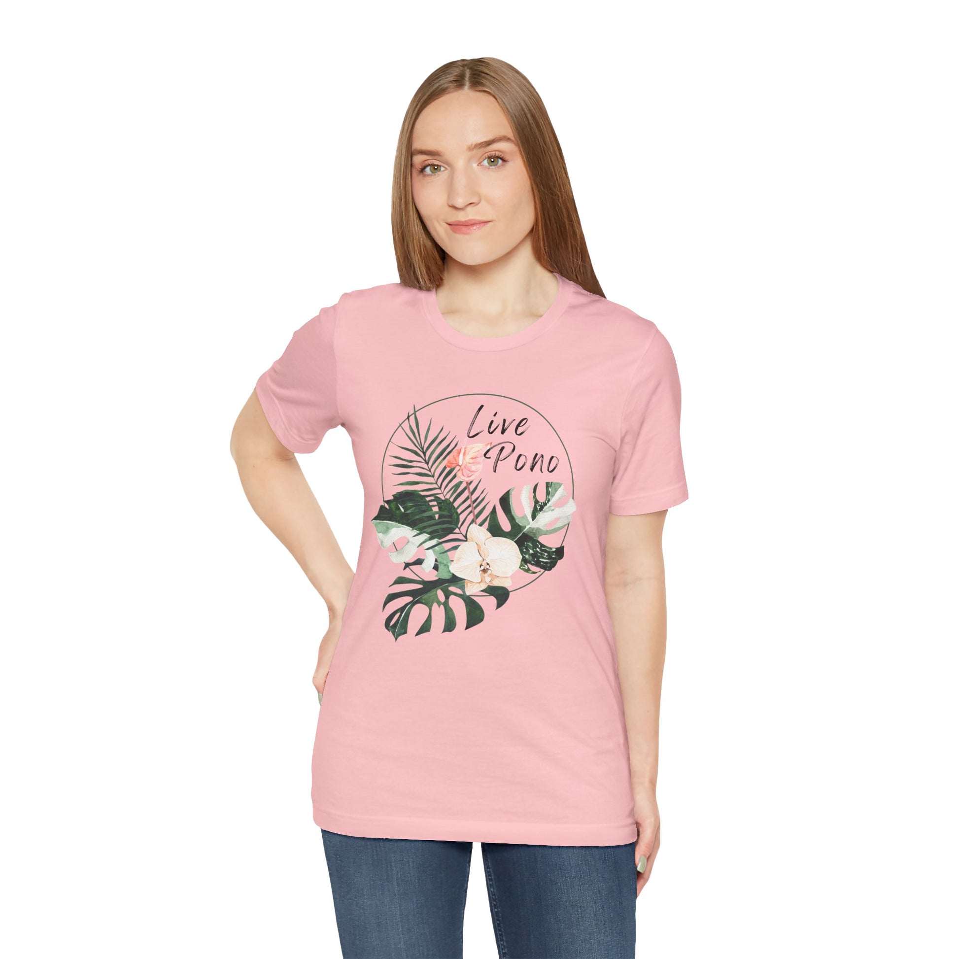 A woman modeling a light pink crewneck t-shirt with a tropical graphic on the front. The graphic features green palm and monstera leaves, white flowers, and the text 'Live in Puno' in black and coral.