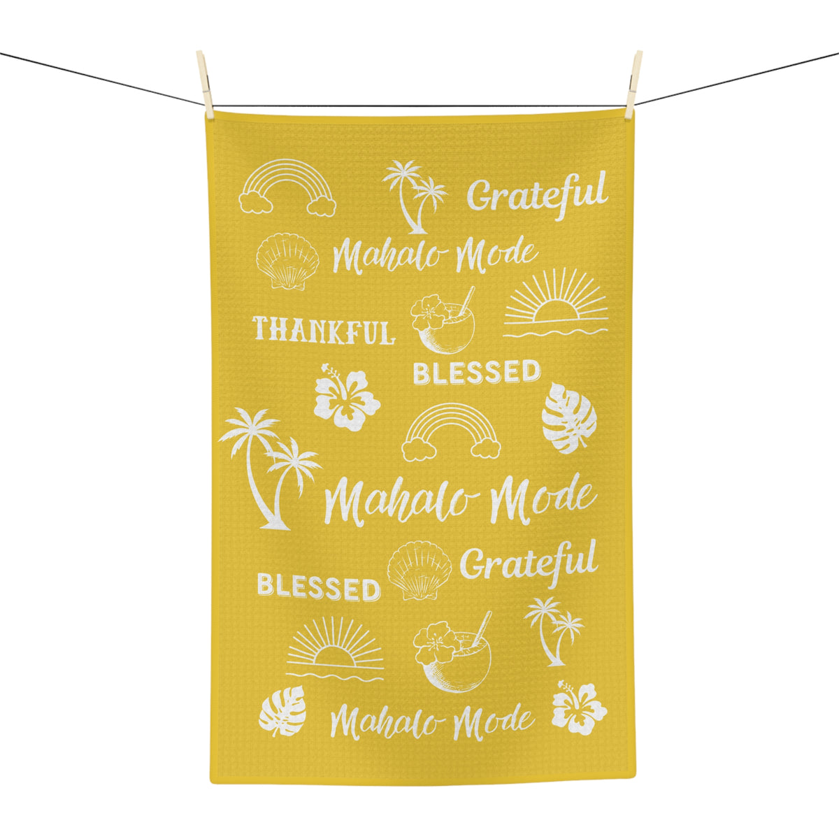 A yellow waffle-weave kitchen towel with white tropical-themed text 'mahalo mode' 'thankful' 'blessed' 'grateful' and illustrations, hanging from a clothesline with wooden clips against a white background.