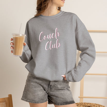 A woman wearing a light grey crewneck sweatshirt with 'Couch Club' printed in pink script across the chest, paired with gray denim shorts and holding an iced coffee.