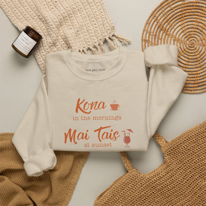 A beige crewneck sweatshirt folded neatly on natural accessoires featuring orange text that reads 'Kona in the mornings Mai Tais at sunset' with small coffee cup and cocktail glass illustrations.