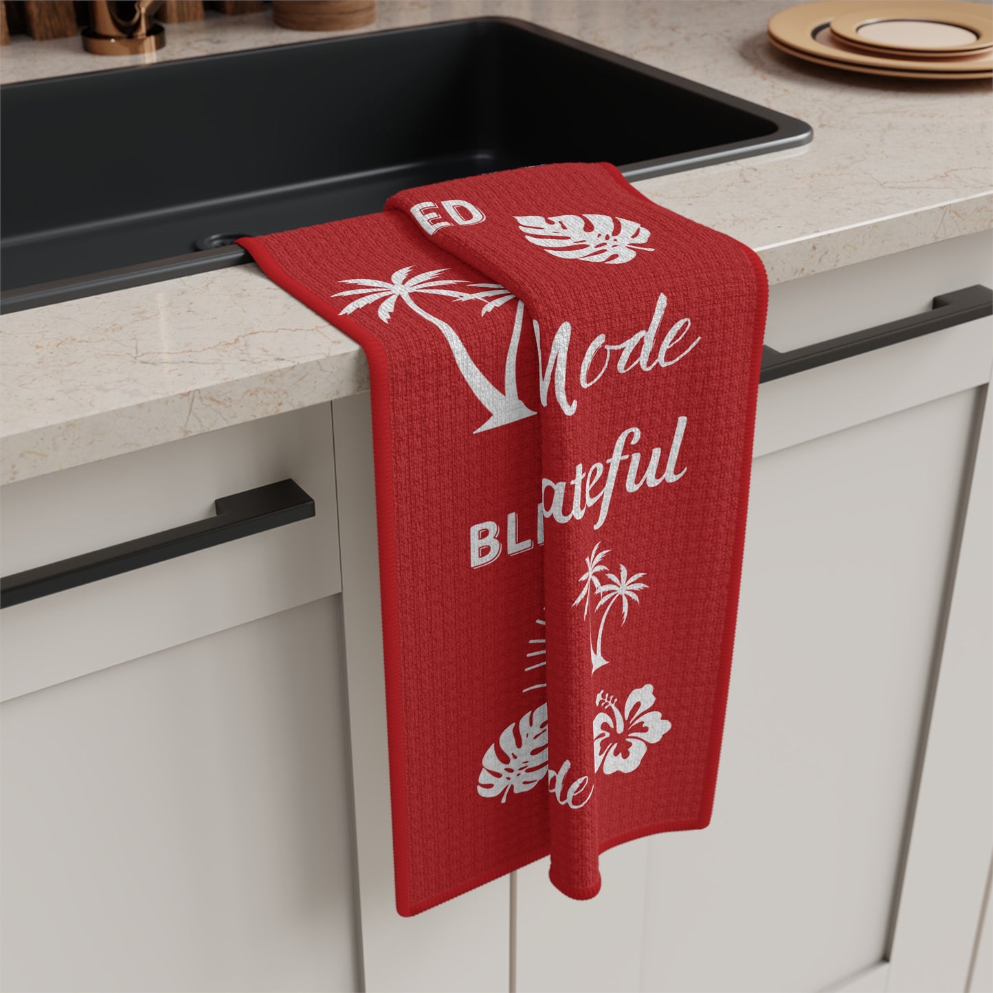 A red waffle-weave kitchen towel with white tropical and text graphics hangs over the edge of a black sink in a light beige countertop kitchen setting.