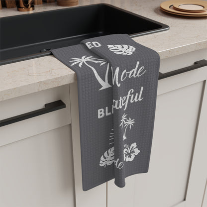 A dark gray kitchen towel with white tropical prints and text hangs over the edge of a black sink in a beige countertop kitchen.