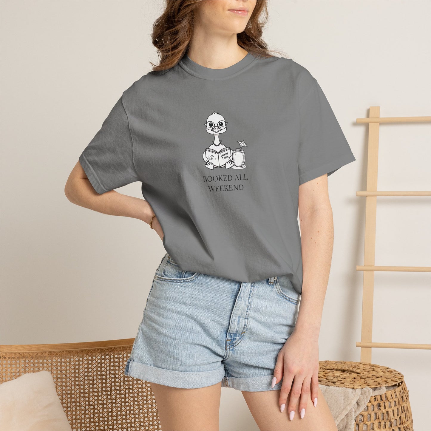 A woman standing wearing a grey comfort colors t-shirt with design of a nene goose reading a book and text 'booked all weekend' paired with denim shorts