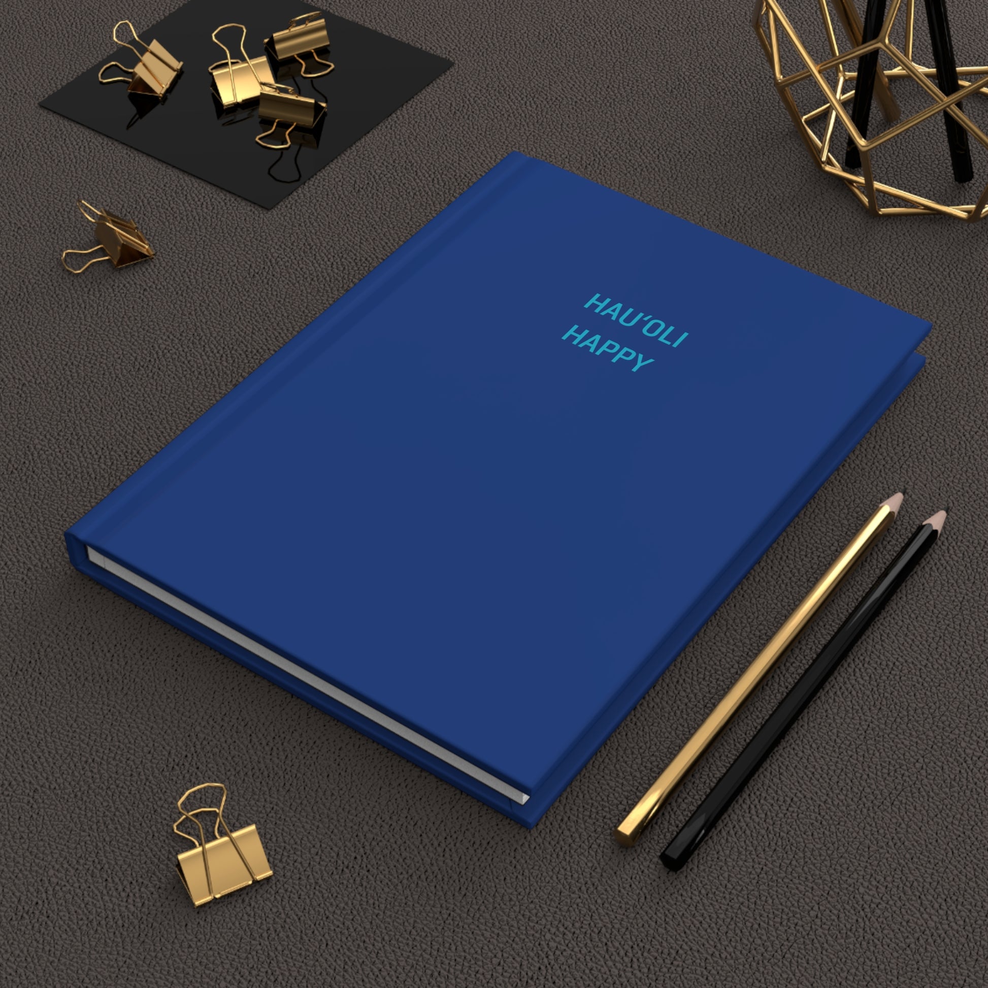 A blue hardcover notebook with the words 'HAUOLI HAPPY' printed in light blue on the cover, styled on a dark textured surface with gold and black office accessories.