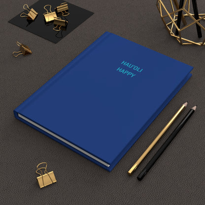 A blue hardcover notebook with the words 'HAUOLI HAPPY' printed in light blue on the cover, styled on a dark textured surface with gold and black office accessories.
