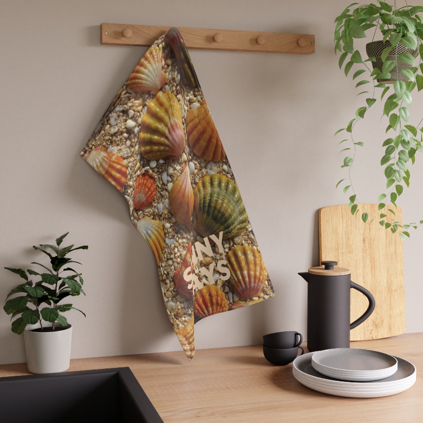 A kitchen towel featuring a realistic print of orange and green sunrise sea shells on a bed of pebbles, hanging from a wooden peg on a beige wall above a kitchen counter with a coffee pot, plates, and plants.