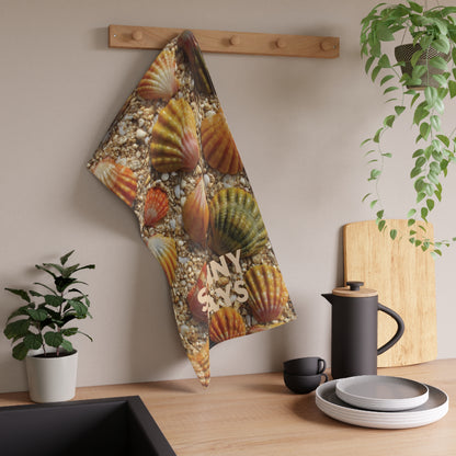 A kitchen towel featuring a realistic print of orange and green sunrise sea shells on a bed of pebbles, hanging from a wooden peg on a beige wall above a kitchen counter with a coffee pot, plates, and plants.