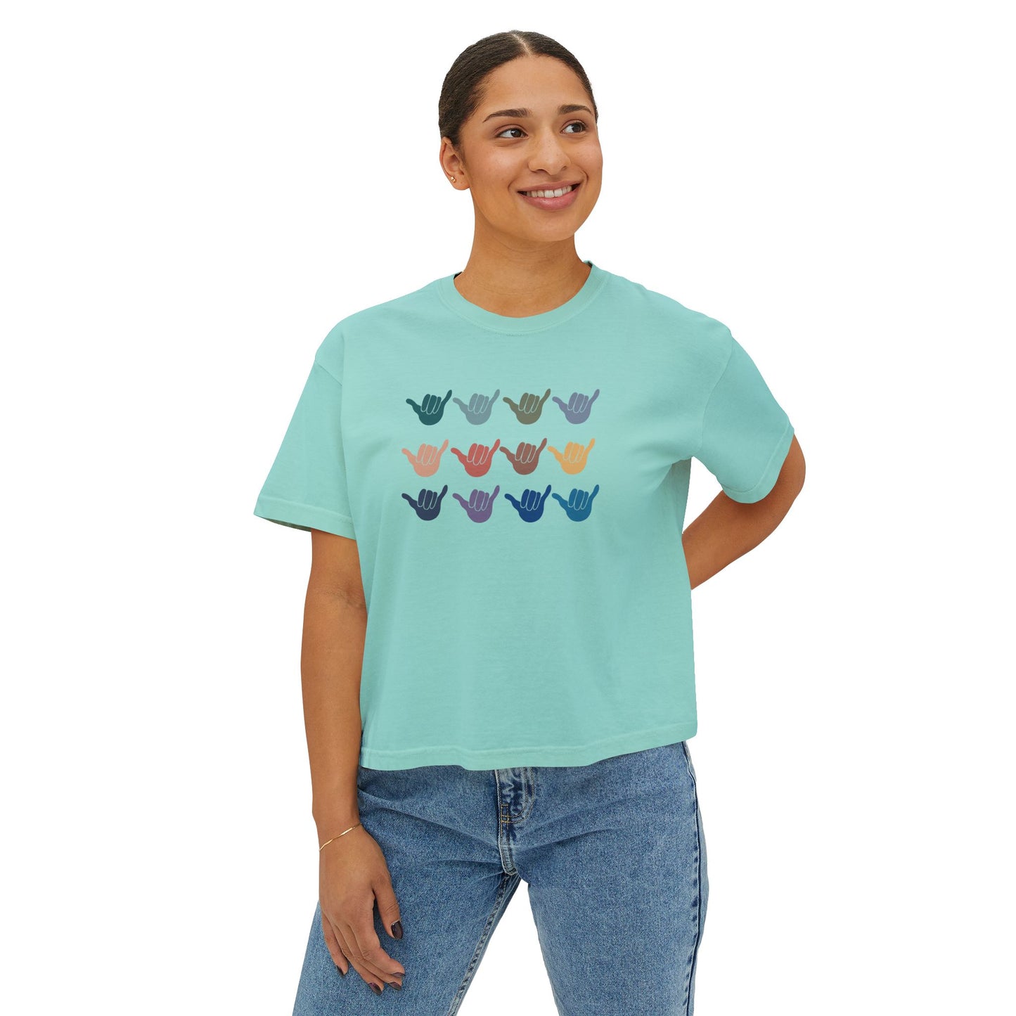 A woman wearing a light mint green short-sleeve crop top with 12 shakas graphic on the front paired with blue jeans, against a plain white background.