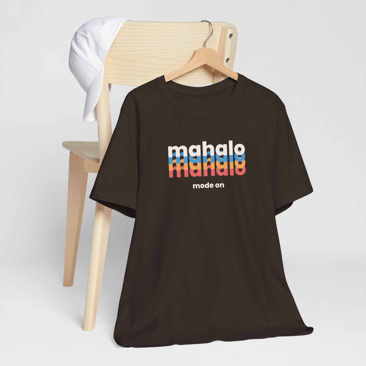 A dark brown short-sleeve t-shirt on a wooden hanger, styled in front of a light wood chair with a white cap draped over the backrest. The shirt features a retro-style graphic on the chest reading 'mahalo mode' in white and rainbow stripes, with 'mode on' printed below.