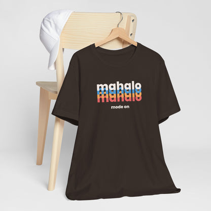 A dark brown short-sleeve t-shirt on a wooden hanger, styled in front of a light wood chair with a white cap draped over the backrest. The shirt features a retro-style graphic on the chest reading 'mahalo mode' in white and rainbow stripes, with 'mode on' printed below.