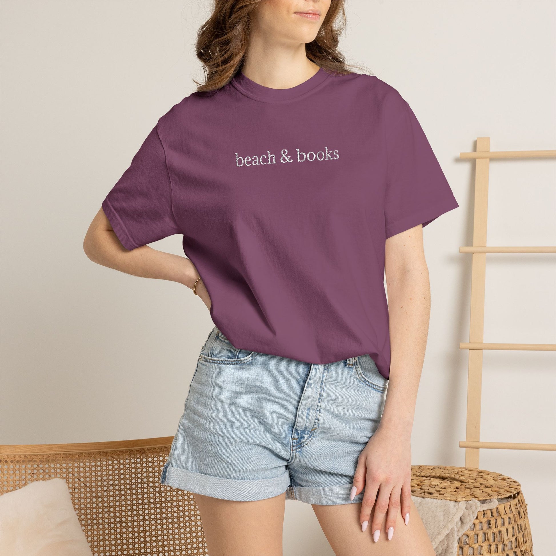 Person wearing a purple comfort colors t-shirt with embroidered text 'beach & books' and denim shorts on a neutral background