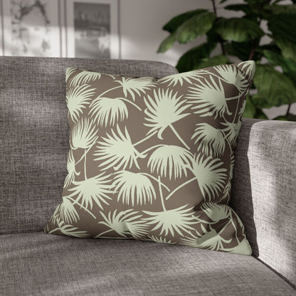 A square throw pillow with a light sage green palm leaf pattern on a taupe-brown background, resting on a gray textured sofa with a blurred indoor plant in the background.