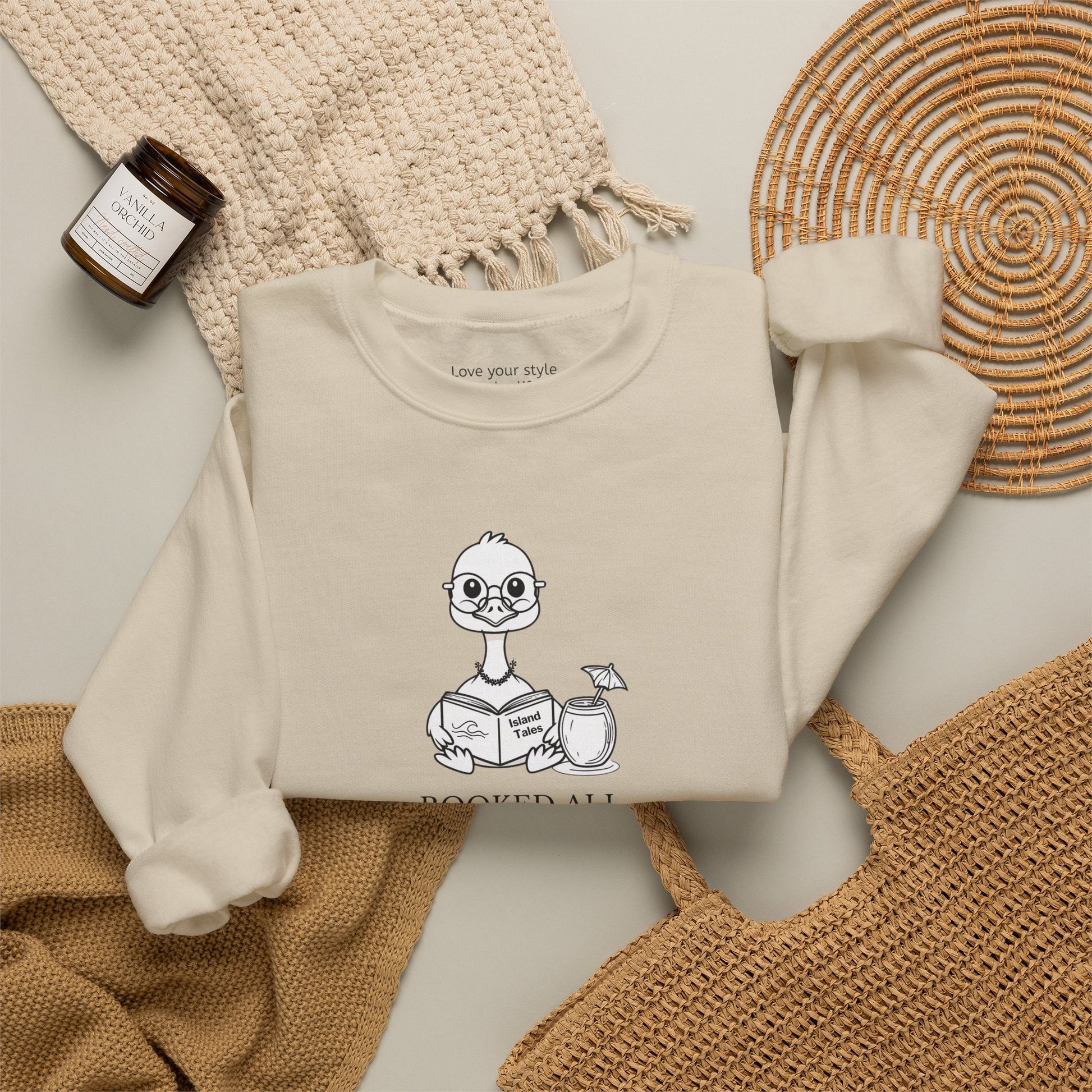 A sand sweatshirt laid flat on a neutral surface, featuring a cartoon nene goose reading a book and the text 'booked all weekend' on the front. The sweatshirt is styled with a beige knit blanket, a woven straw mat, a small candle jar, and a tan tote bag.