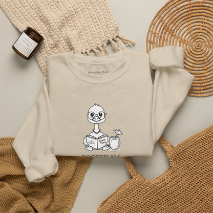 A sand sweatshirt laid flat on a neutral surface, featuring a cartoon nene goose reading a book and the text 'booked all weekend' on the front. The sweatshirt is styled with a beige knit blanket, a woven straw mat, a small candle jar, and a tan tote bag.