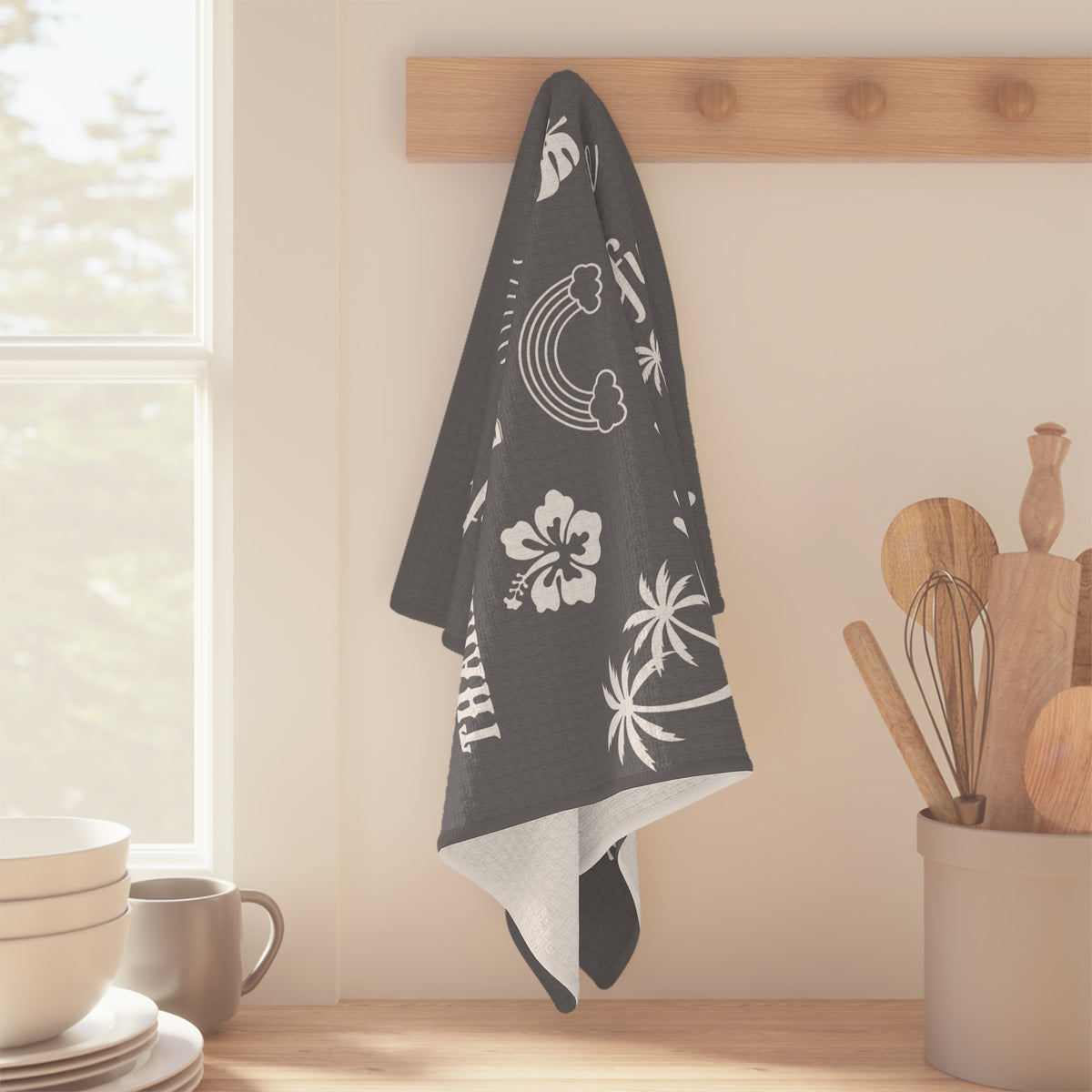 A dark gray kitchen towel with white tropical and Hawaiian prints hangs from a wooden peg on a wall in a bright kitchen setting.