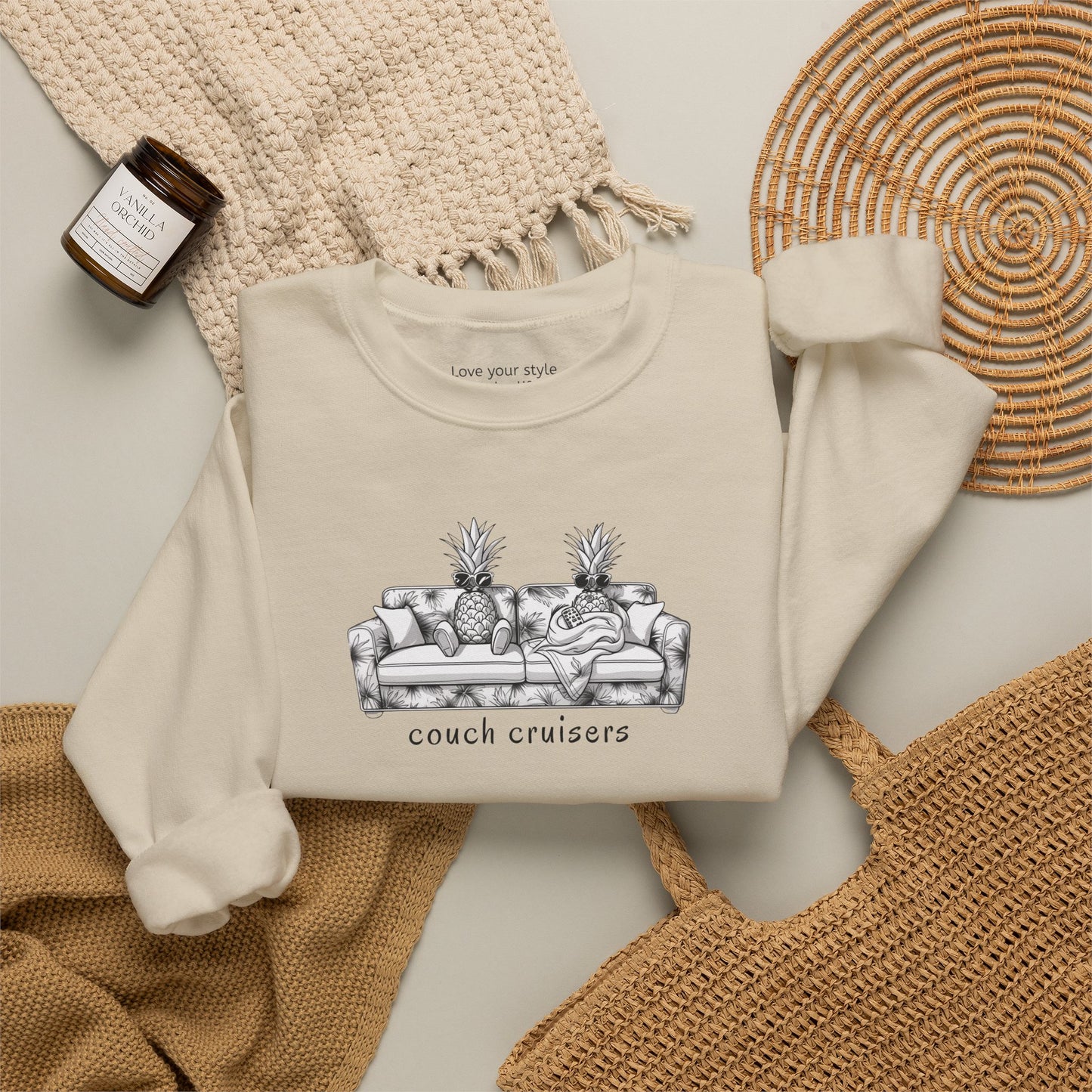 A sand-colored crewneck sweatshirt with a black and white graphic of a couch with pineapples and the text 'couch cruisers', styled in a flat lay with a woven blanket, a candle, and a wicker tray.