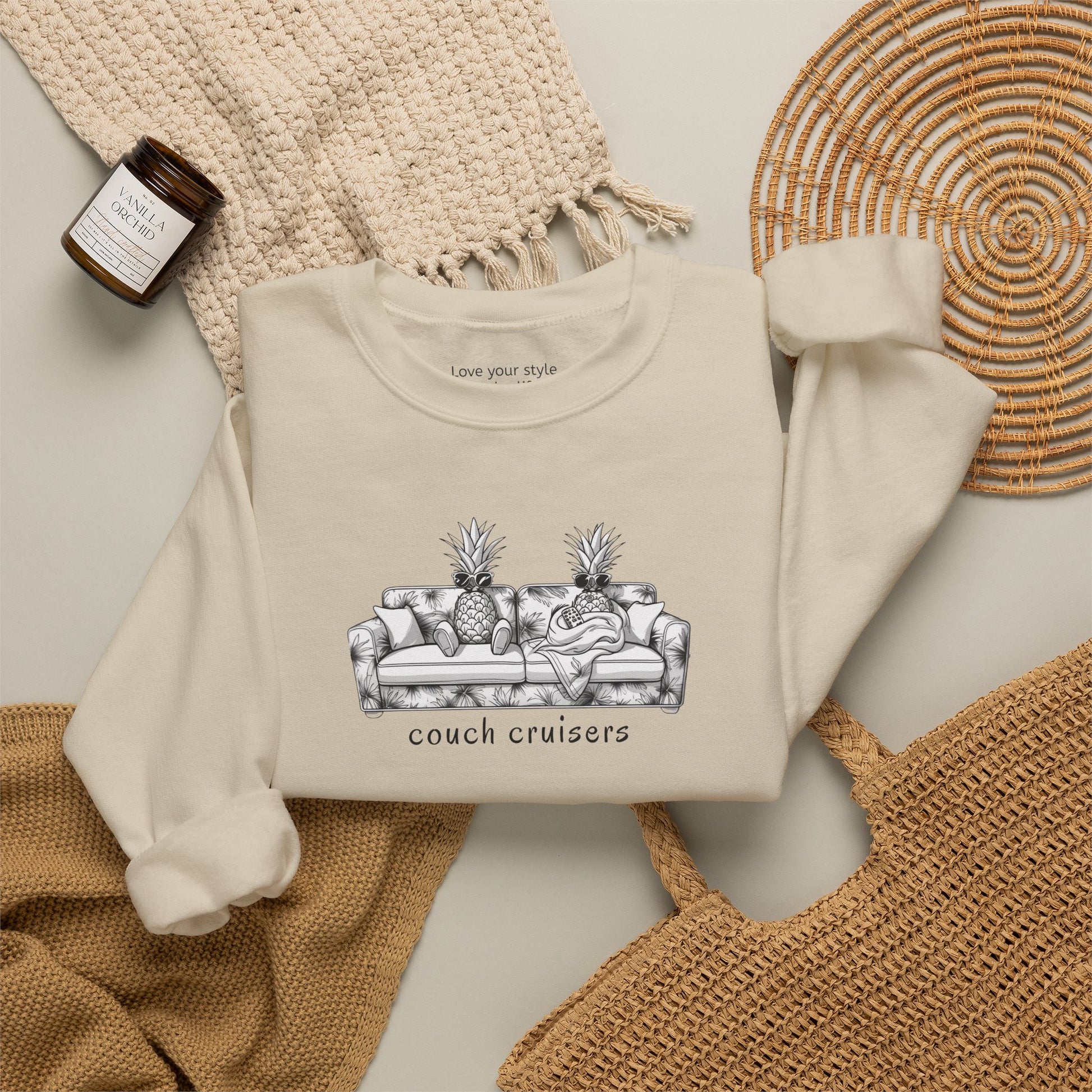 A sand-colored crewneck sweatshirt with a black and white graphic of a couch with pineapples and the text 'couch cruisers', styled in a flat lay with a woven blanket, a candle, and a wicker tray.