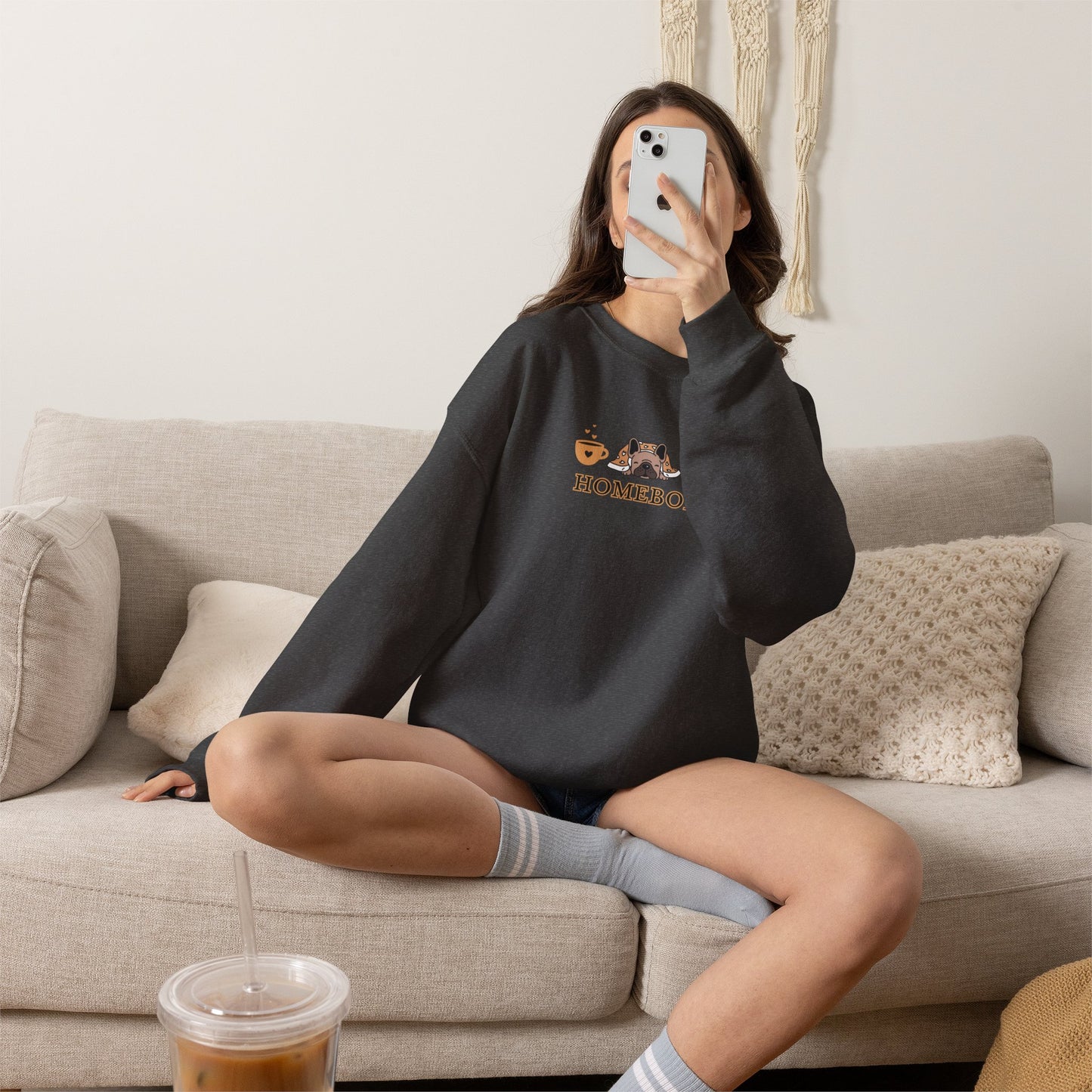 Person wearing a dark grey crewneck sweatshirt featuring a central graphic of a dog under a blanket, house slippers, coffee cups and the word 'HOMEBODY' printed below it sitting on a couch taking a picture.