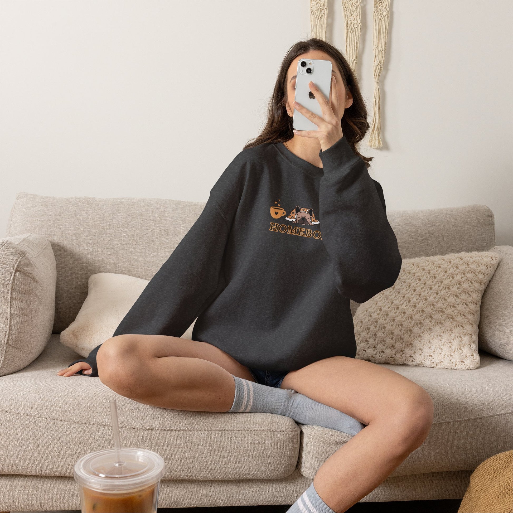 Person wearing a dark grey crewneck sweatshirt featuring a central graphic of a dog under a blanket, house slippers, coffee cups and the word 'HOMEBODY' printed below it sitting on a couch taking a picture.