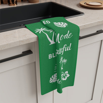 A green kitchen towel with white tropical and text graphics hanging over the edge of a black sink in a kitchen setting.