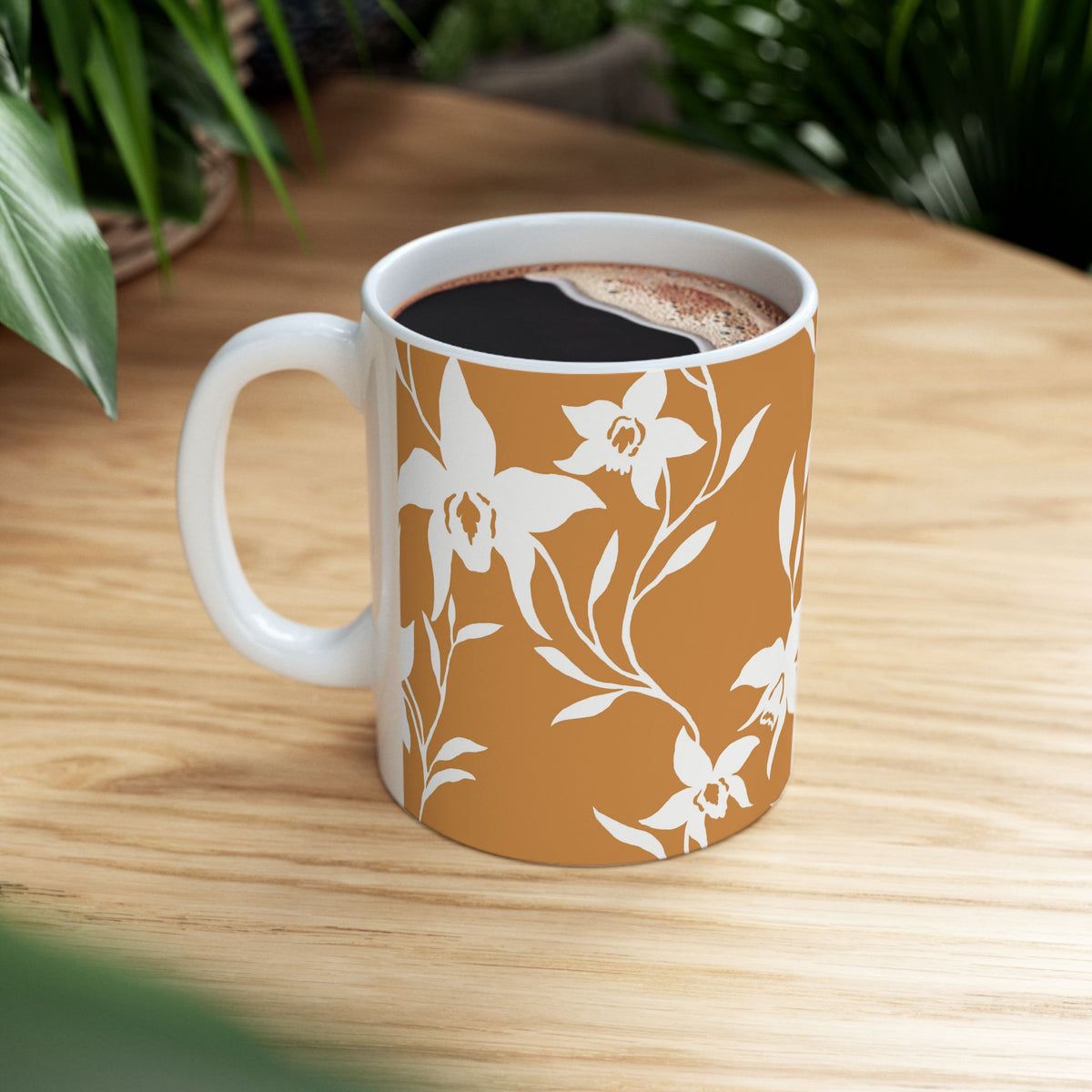 A ceramic coffee mug with a white floral pattern on a terracotta orange background, filled with black coffee and sitting on a light wood table with green plants in the background.