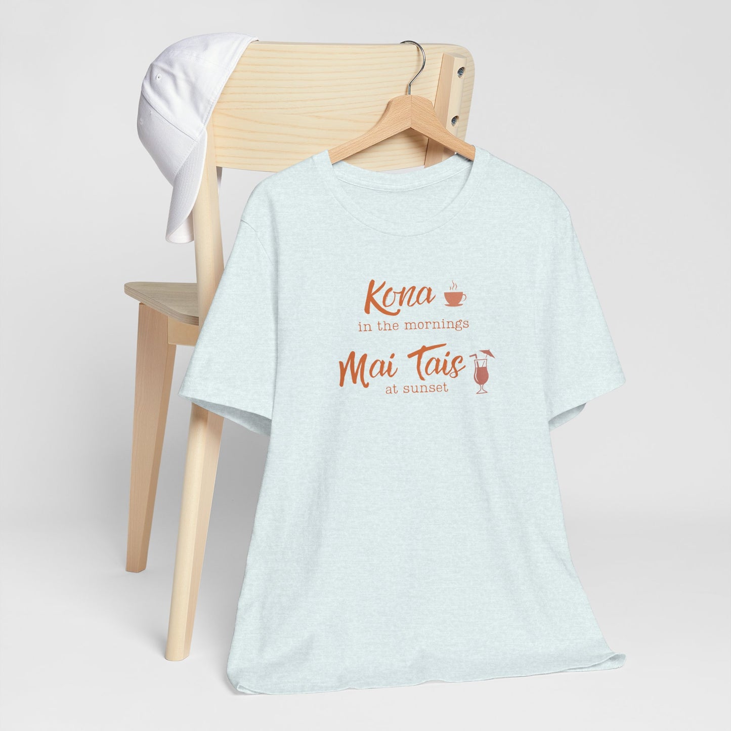 A light heather blue t-shirt hanging on a wooden hanger from a wooden chair, featuring a coral orange graphic that reads 'Kona in the mornings Mai Tais at sunset' with small coffee and cocktail illustrations.