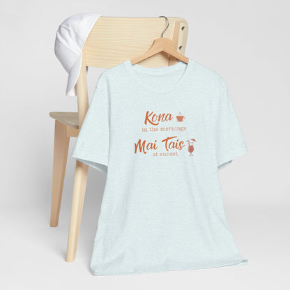 A light heather blue t-shirt hanging on a wooden hanger from a wooden chair, featuring a coral orange graphic that reads 'Kona in the mornings Mai Tais at sunset' with small coffee and cocktail illustrations.