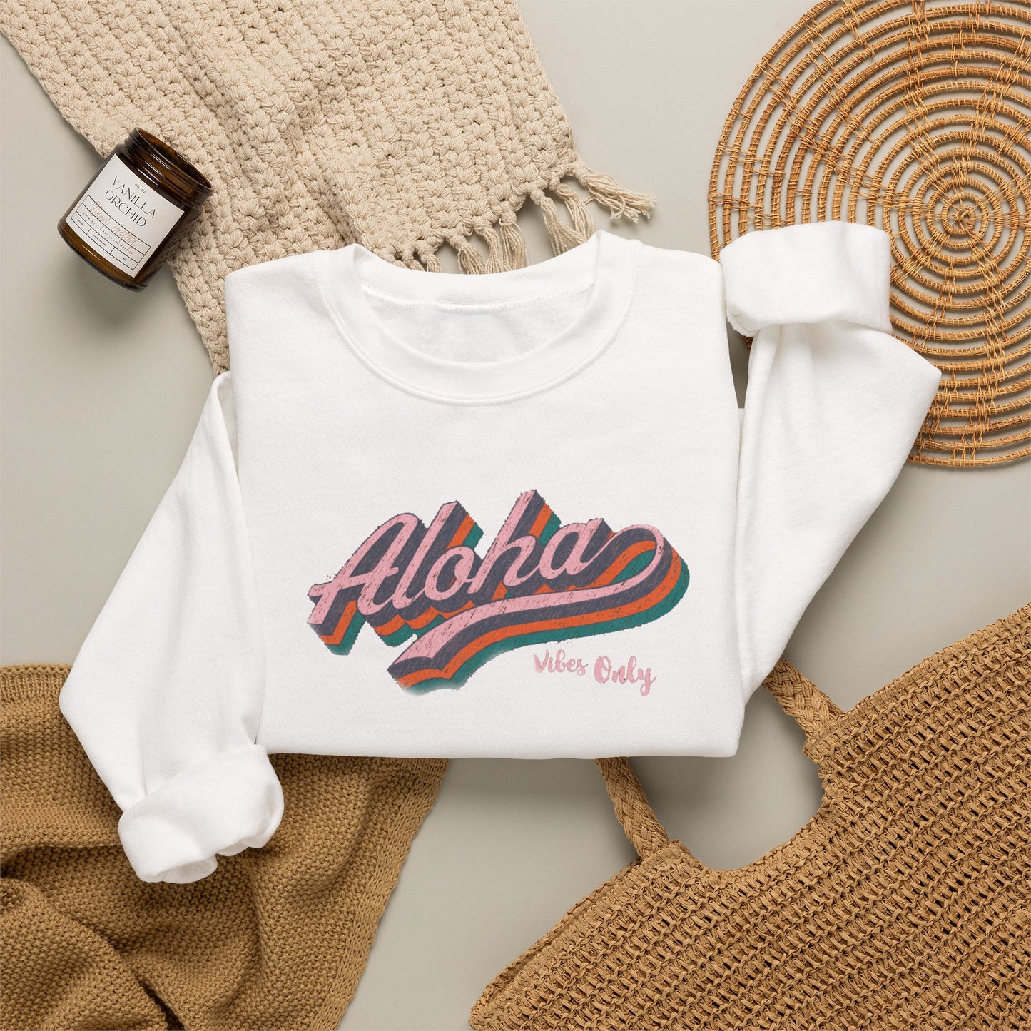 A folded white long-sleeve sweatshirt with a colorful retro-style 'Aloha Vibes Only' graphic, styled in a flat lay with a beige knit blanket, a woven mat, a small candle jar, and a tan woven bag on a light gray surface.