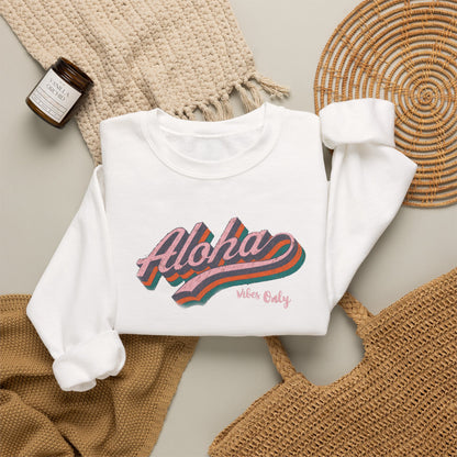 A folded white long-sleeve sweatshirt with a colorful retro-style 'Aloha Vibes Only' graphic, styled in a flat lay with a beige knit blanket, a woven mat, a small candle jar, and a tan woven bag on a light gray surface.