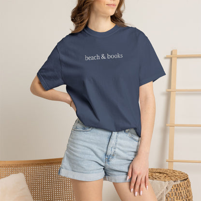 Person wearing a blue comfort colors t-shirt with embroidered text 'beach & books' and denim shorts on a neutral background