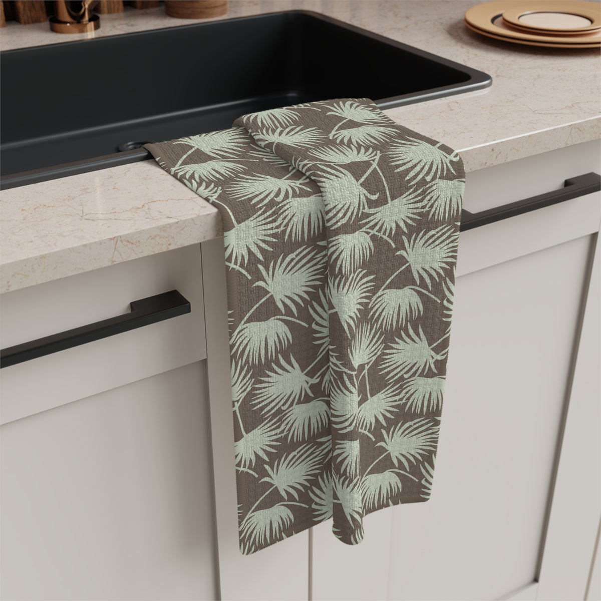 A kitchen towel with a light green palm leaf pattern on a taupe background, draped over the edge of a black sink in a modern kitchen setting.