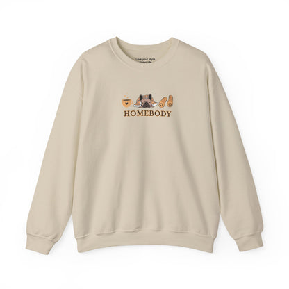 A beige crewneck sweatshirt featuring a central graphic of a dog under a blanket, house slippers, coffee cups and the word 'HOMEBODY' printed below it.